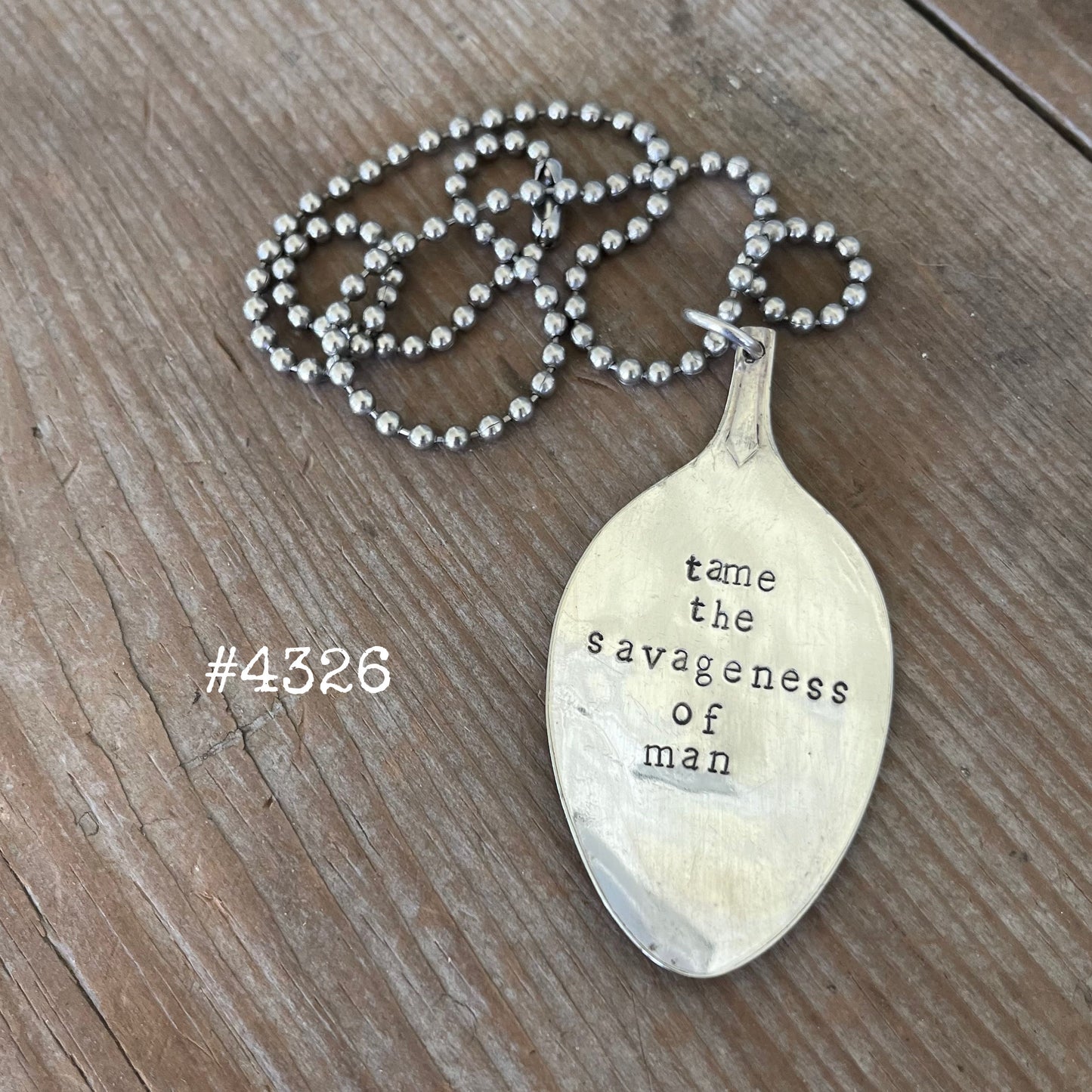 Close-up of hand-stamped spoon pendant with 'Tame the Savageness of Man' quote showing reclaimed vintage silverware, one-of-a-kind compassion-themed design