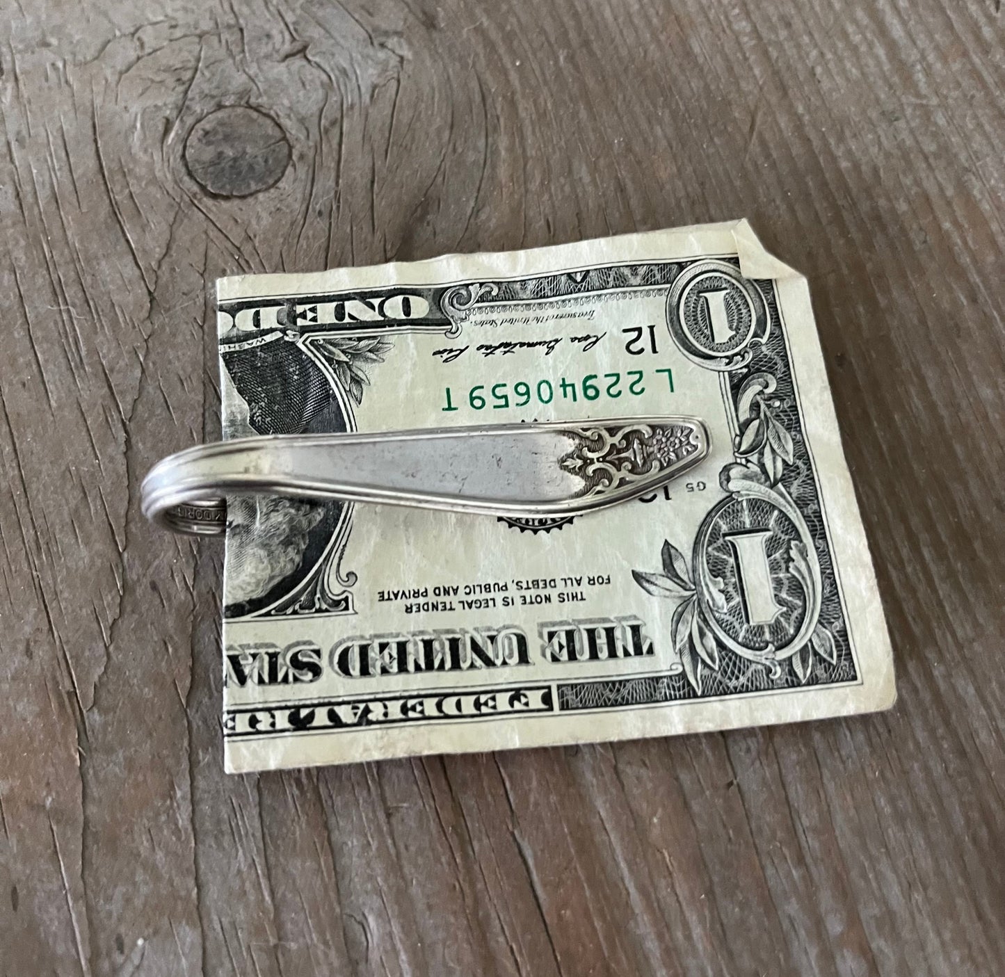 Handle view of a vintage silver-plate sugar spoon money clip with a design resembling a guitar on the back, resting on a dollar bill.