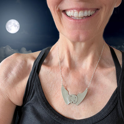 Bat pendant made from upcycled vintage spoon bowls shown on model with full moon in the back - handmade Halloween artisan necklace - handcrafted upcycled vintage silverware jewelry - Laughing Frog Studio