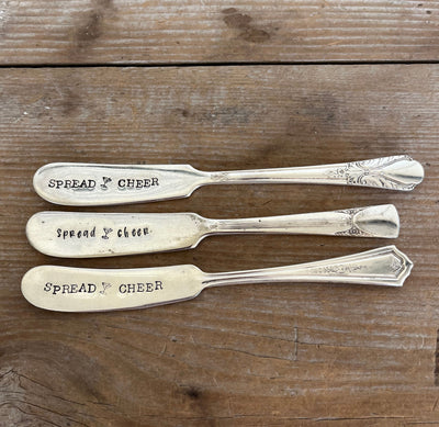 Three "Spread Cheer" hand-stamped cheese spreaders comparison flat lay on wood - handcrafted from upcycled vintage silverware - Laughing Frog Studio