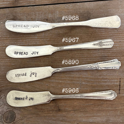 Close up of Side view of four "Spread Joy" hand-stamped cheese spreaders comparison flat lay on wood - hand-stamped cheese spreader - handcrafted from upcycled vintage silverware - Laughing Frog Studio