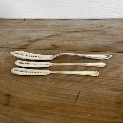 Side view of three "Who Cut the Cheese?" hand-stamped cheese spreaders comparison flat lay on wood - handcrafted from upcycled vintage silverware - Laughing Frog Studio