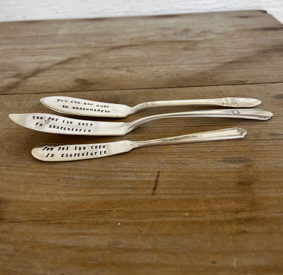 Side view of three "You Put the Cute in Charcuterie" hand-stamped cheese spreaders flat lay on wood - handcrafted from upcycled vintage silverware - Laughing Frog Studio