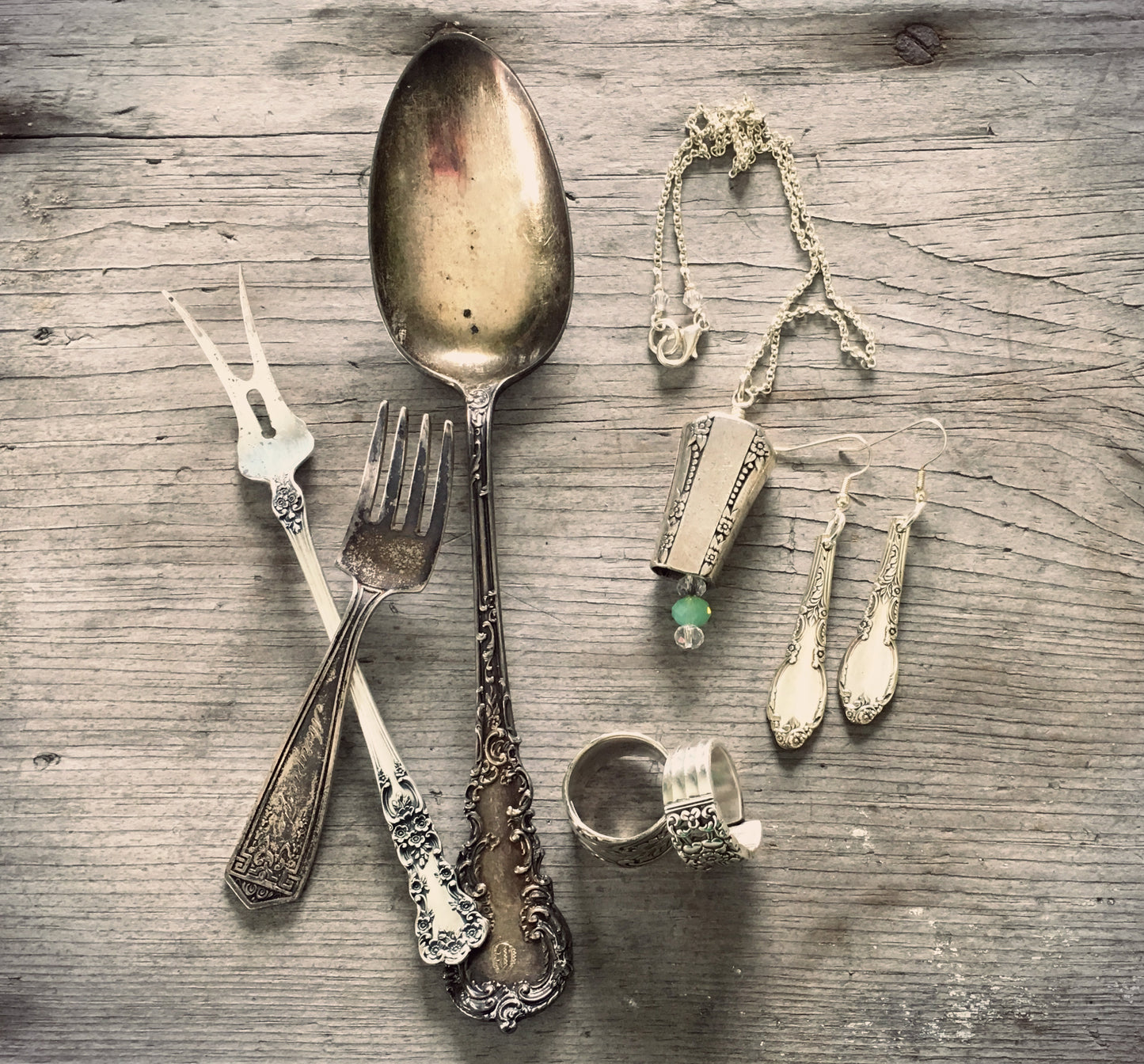 Ornate vintage spoons and forks laid next to upcycled spoon ring, earrings, and necklace — handcrafted silverware jewelry flat lay