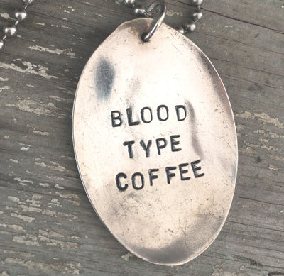 Close-up of BLOOD TYPE COFFEE hand-stamped spoon pendant showing bold lettering on reclaimed vintage silverware, caffeine-themed design