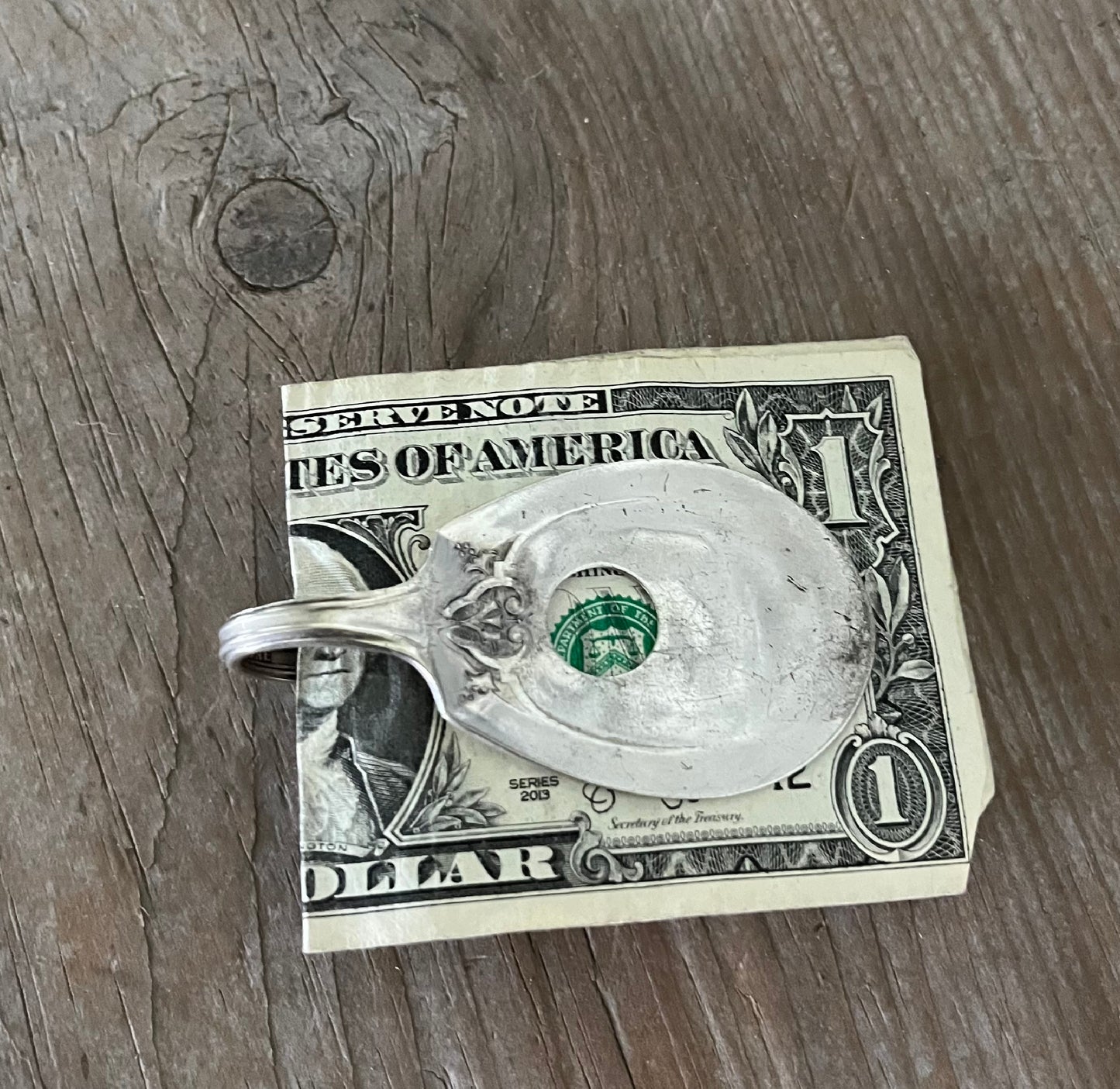 A vintage silver-plate sugar spoon money clip with a design resembling a guitar on the back, resting on a dollar bill.