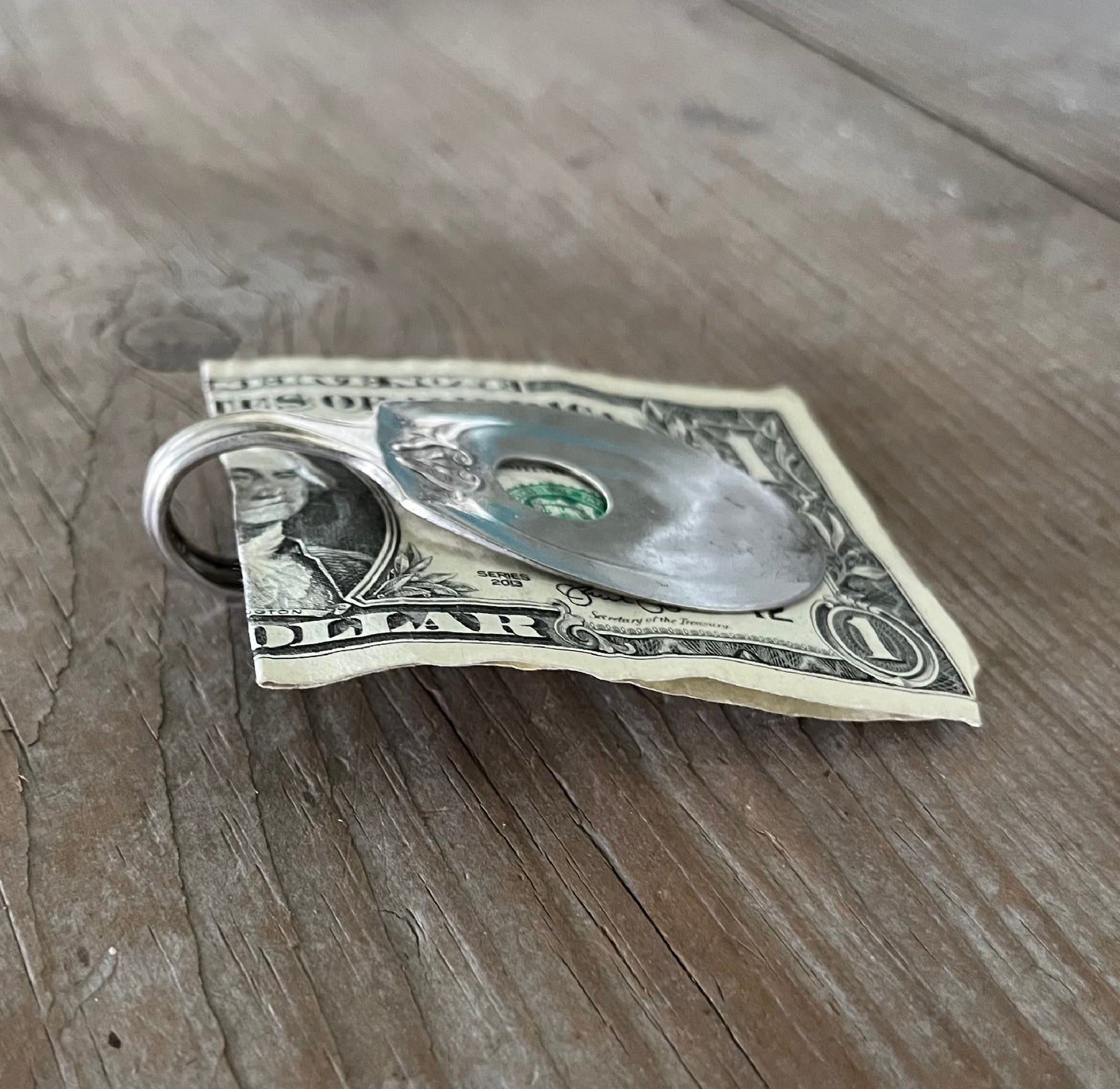Side view of A vintage silver-plate sugar spoon money clip with a design resembling a guitar on the back, resting on a dollar bill.
