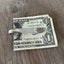 Handle view of a vintage silver-plate sugar spoon money clip with a design resembling a guitar on the back, resting on a dollar bill.