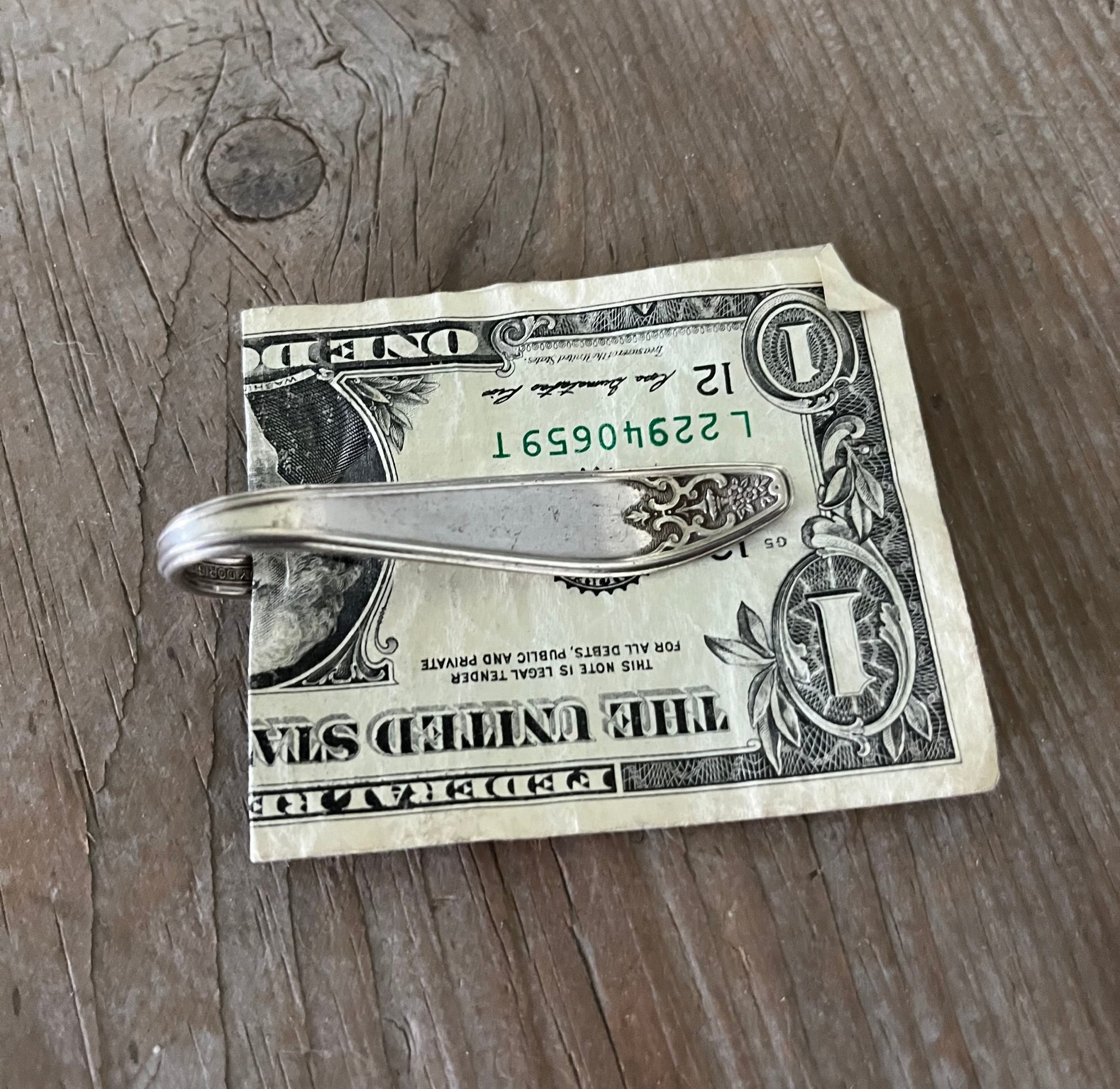 Handle view of a vintage silver-plate sugar spoon money clip with a design resembling a guitar on the back, resting on a dollar bill.