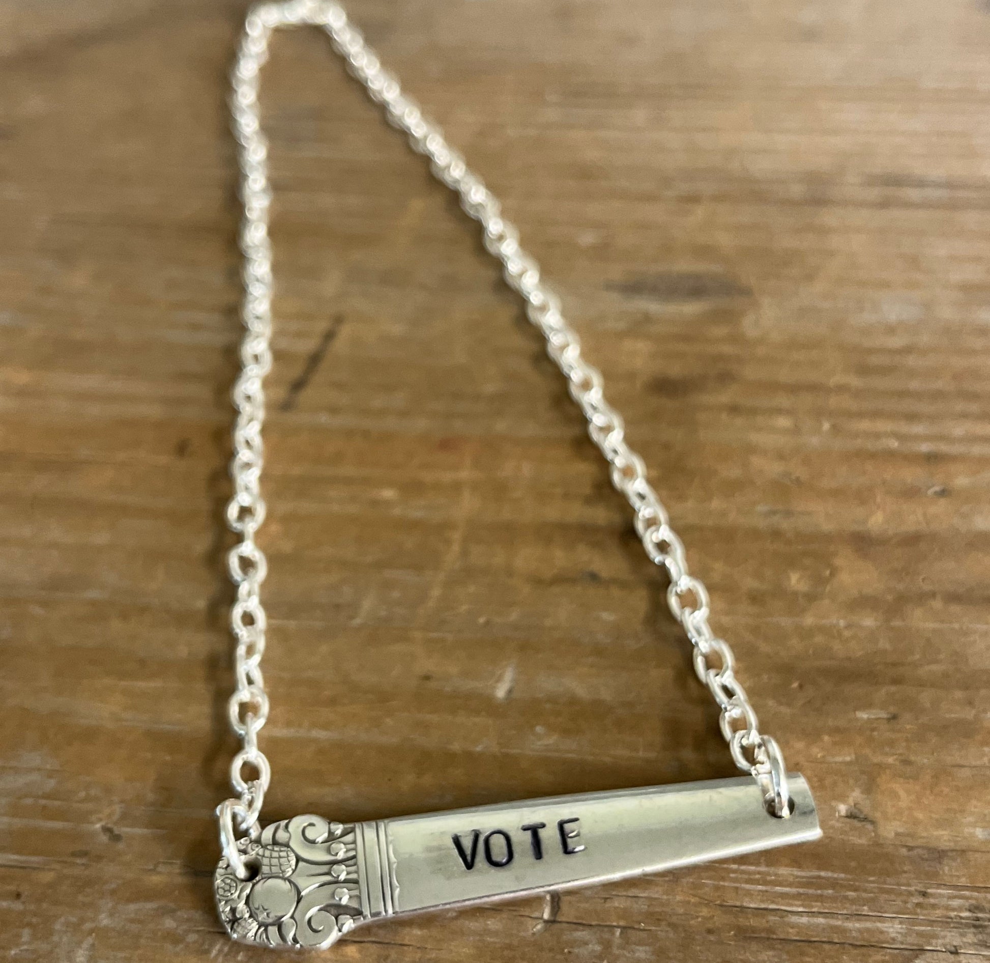 Flat lay of hand-stamped VOTE bar choker necklace showing full silver chain and vintage silverware handle pendant with floral graphic detail - handcrafted from upcycled vintage silverware - Laughing Frog Studio