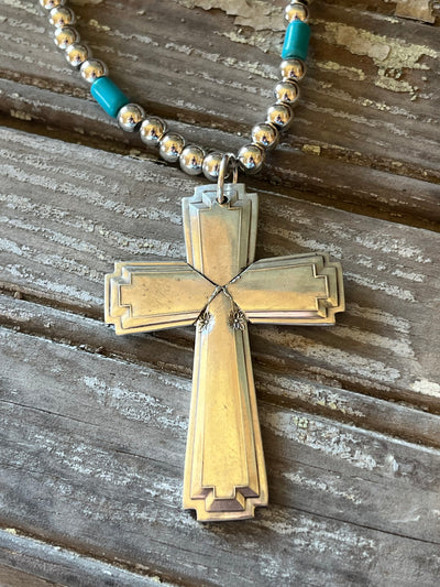Spoon cross pendant on a silverplate bead choker with turquoise-colored accent beads, resting on weathered wood.