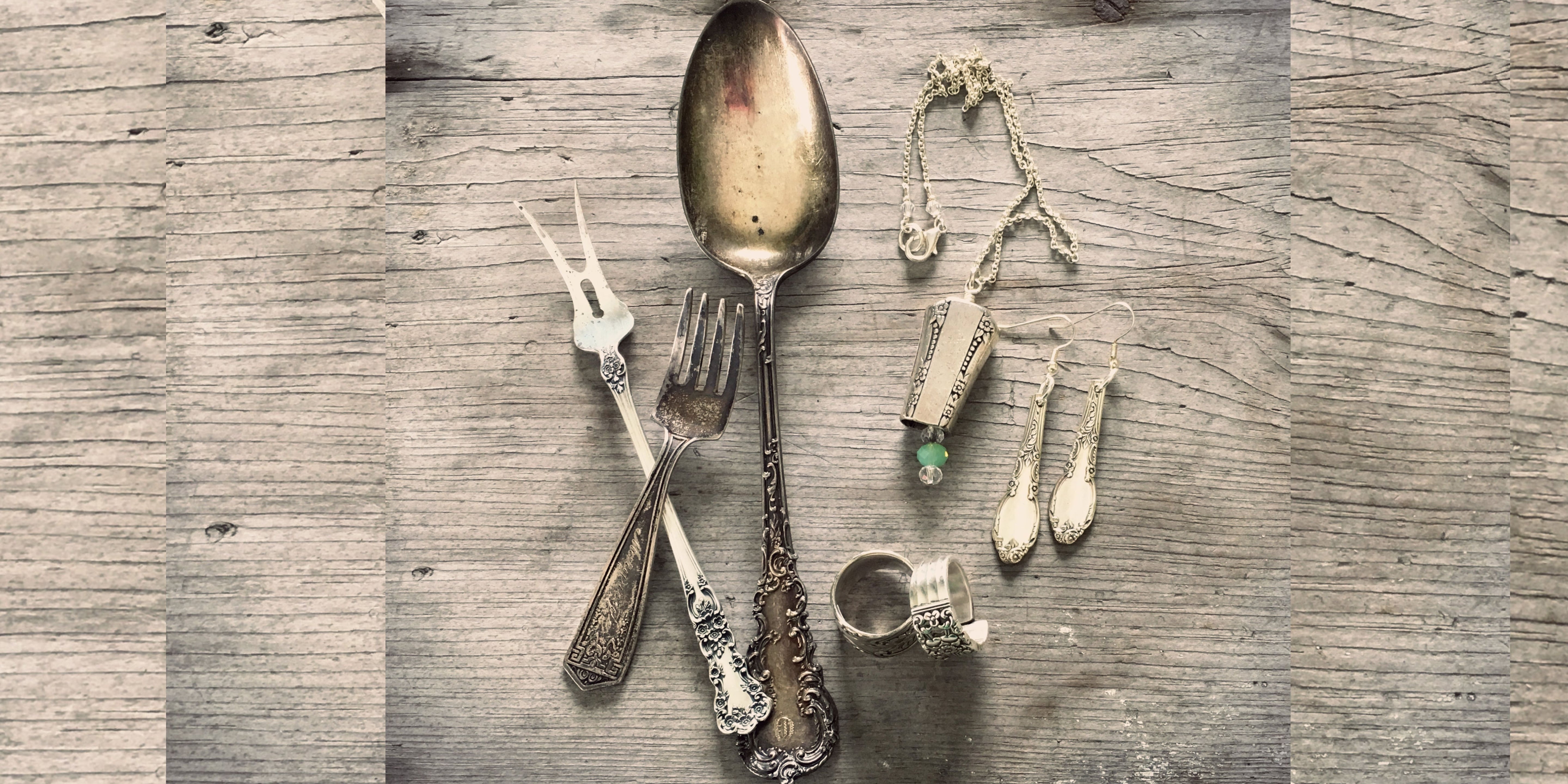 Flat lay of vintage silverware alongside handcrafted jewelry on weathered wood — spoon, fork, and pickle fork with upcycled spoon ring, earrings, and necklace — Laughing Frog Studio