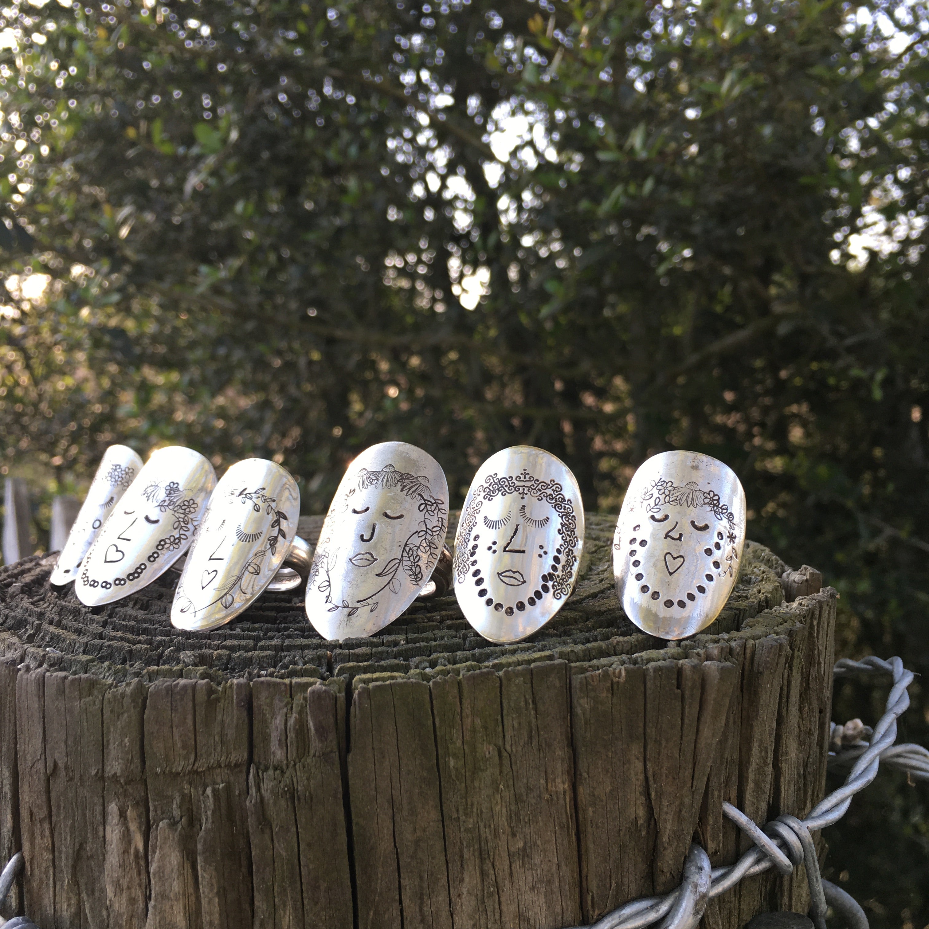 Five Women of Nature hand-stamped spoon rings displayed on a weathered fence post outdoors — upcycled vintage silverware rings