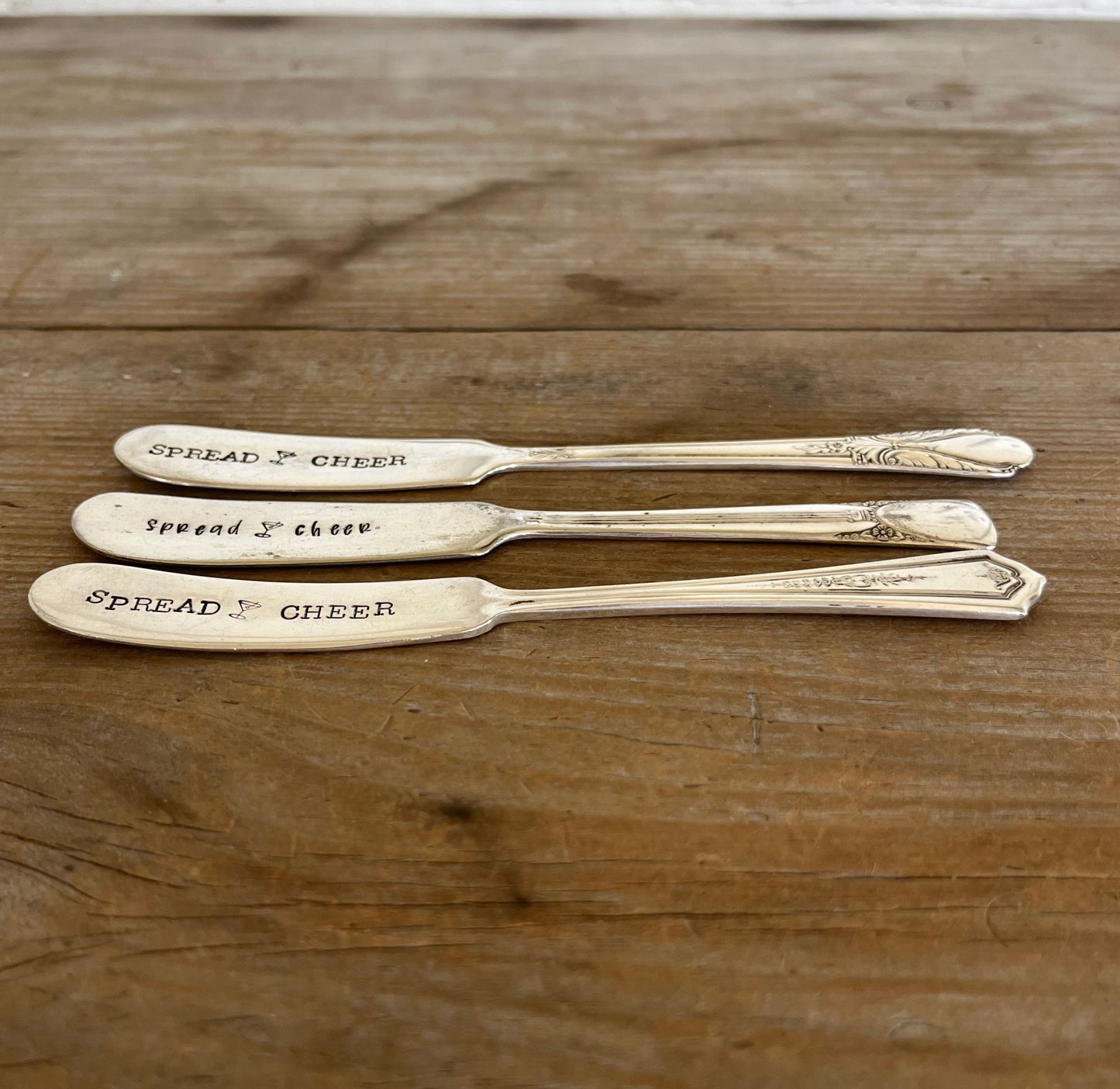 Side view of three "Spread Cheer" hand-stamped cheese spreaders comparison flat lay on wood - handcrafted from upcycled vintage silverware - Laughing Frog Studio