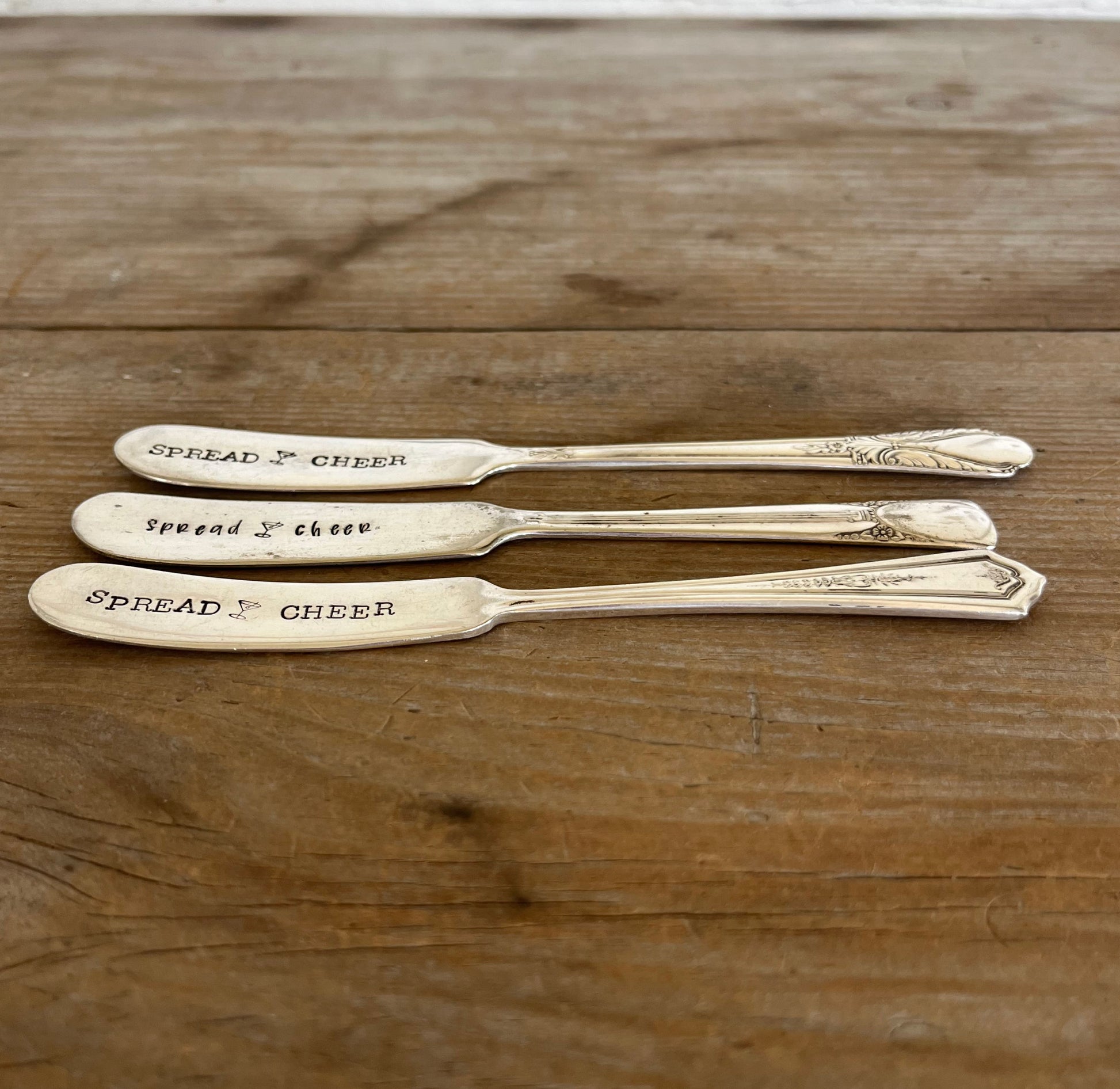 Side view of three "Spread Cheer" hand-stamped cheese spreaders comparison flat lay on wood - handcrafted from upcycled vintage silverware - Laughing Frog Studio