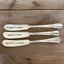 Three "Spread Cheer" hand-stamped cheese spreaders comparison flat lay on wood - handcrafted from upcycled vintage silverware - Laughing Frog Studio