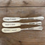 Three "Spread Cheer" hand-stamped cheese spreaders comparison flat lay on wood - hand-stamped cheese spreader - handcrafted from upcycled vintage silverware - Laughing Frog Studio