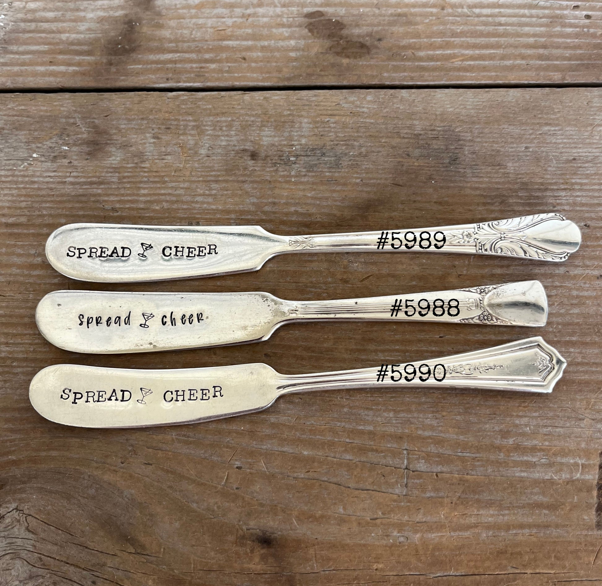 Three "Spread Cheer" hand-stamped cheese spreaders comparison flat lay on wood - hand-stamped cheese spreader - handcrafted from upcycled vintage silverware - Laughing Frog Studio