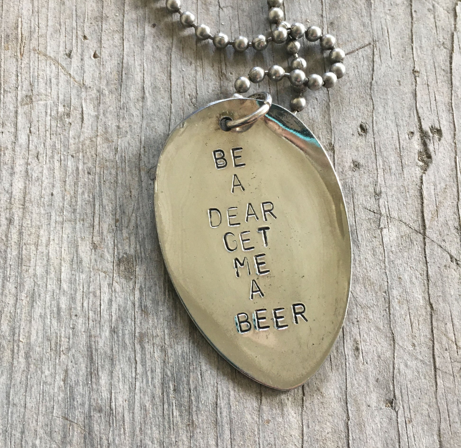 Close-up of BE A DEAR GET ME A BEER hand-stamped spoon pendant showing cheeky message on reclaimed vintage silverware, feminist humor design