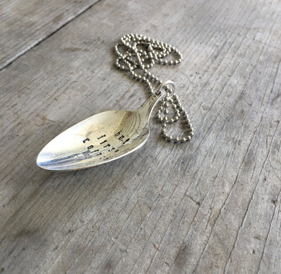 Close-up of BUT FIRST COFFEE hand-stamped spoon pendant with steaming cup showing quirky message on reclaimed vintage silverware, 18-inch chain